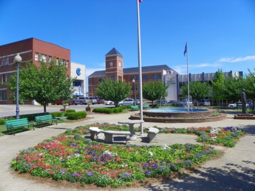 A vibrant park with colorful flowers, benches, and a fountain, surrounded by buildings under a clear blue sky.
