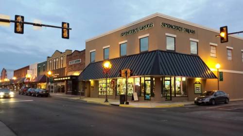 A street view of a corner building with "Spencer's Drugs" signage, illuminated at dusk, surrounded by shops and parked cars.