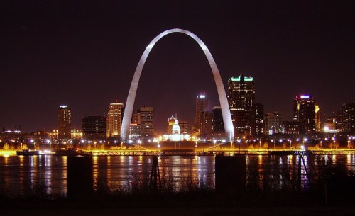 The Gateway Arch illuminated at night, with a city skyline and river reflecting the lights.