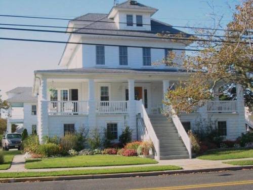A large white house with a porch, surrounded by greenery and flowers, on a sunny day.