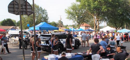 Community event with tents, a police vehicle, and people socializing in a lively outdoor setting.