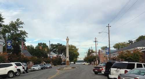 A quiet street scene featuring a statue in the center, lined with trees and parked cars on either side.