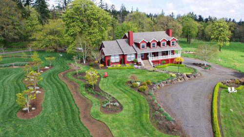 Aerial view of a red house with a porch, surrounded by lush green landscaping and winding garden paths.