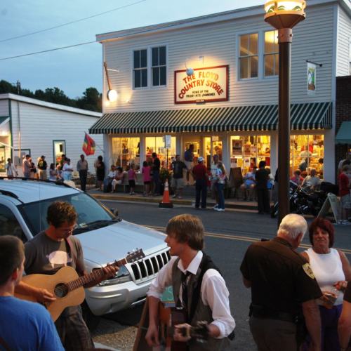 A lively street scene with people gathered outside a country store, musicians playing, and a car parked nearby.