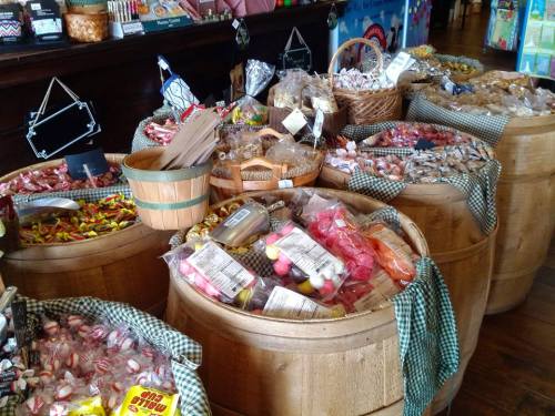 A variety of colorful candies displayed in wooden barrels at a shop, with baskets and checkered cloths.
