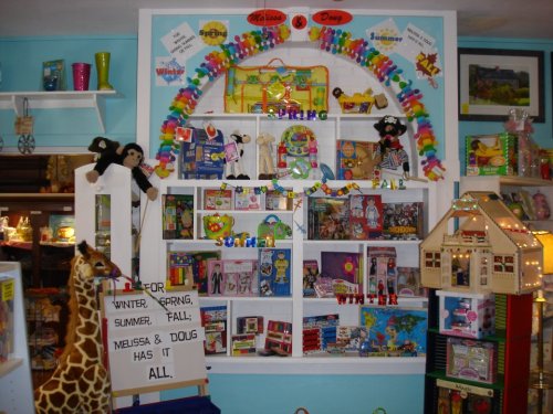 Colorful display of toys and games on shelves, featuring stuffed animals, dolls, and a sign promoting Melissa & Doug products.