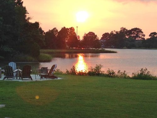 A serene sunset over a lake, with silhouettes of trees and chairs on a grassy shore reflecting in the water.