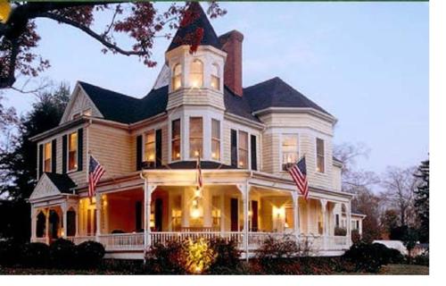 A charming Victorian-style house with a turret, illuminated windows, and American flags on the porch at dusk.