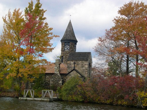A stone church with a clock tower surrounded by colorful autumn trees by a calm waterway.