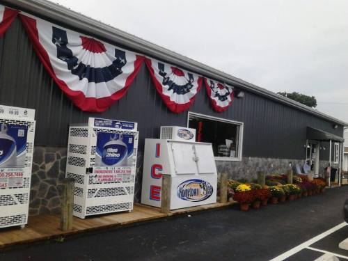 A storefront with patriotic bunting, ice machines, and flower pots, set against a gray building.