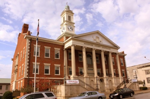 Historic brick courthouse with a clock tower, columns, and an American flag, set against a partly cloudy sky.