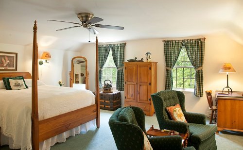 Cozy bedroom with a four-poster bed, green armchairs, wooden furniture, and large windows with checkered curtains.