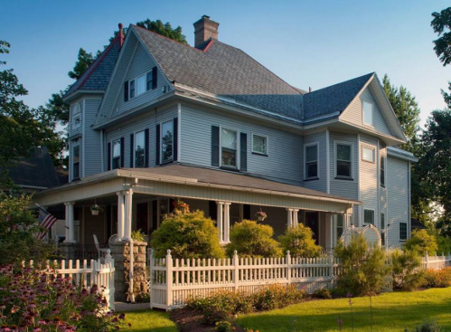 A large, two-story gray house with a porch, surrounded by greenery and a white picket fence.