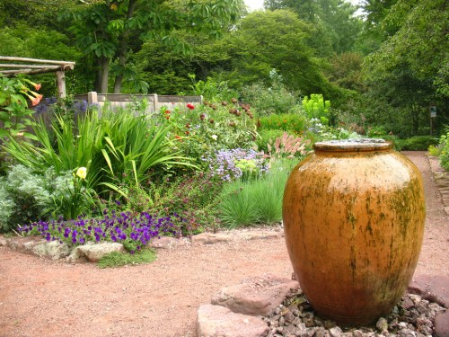 A lush garden with colorful flowers and greenery, featuring a large, weathered clay pot in the foreground.