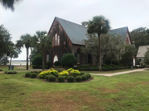A rustic wooden church surrounded by lush greenery and palm trees, near a calm body of water.