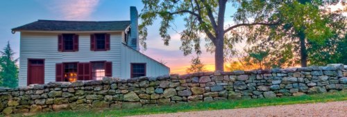 A charming two-story house with red shutters, set against a colorful sunset and a stone wall in the foreground.