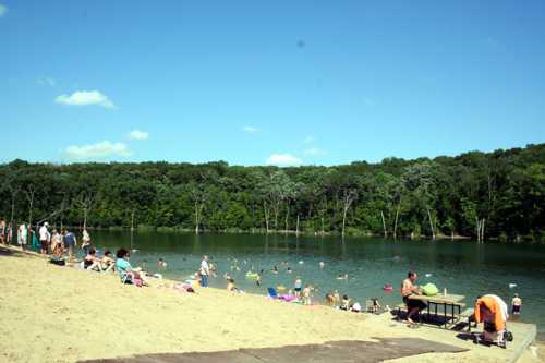 A sandy beach by a lake with people swimming and relaxing under a clear blue sky and green trees in the background.