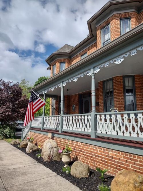A brick house with a porch, American flag, and landscaped garden under a partly cloudy sky.