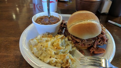 A plate with a pulled pork sandwich, a side of baked beans, and creamy potato salad on a wooden table.