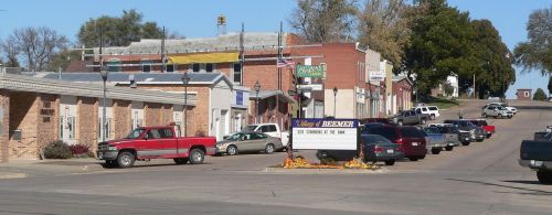 A small town street with parked cars, brick buildings, and a sign welcoming visitors to the village of Bremer.