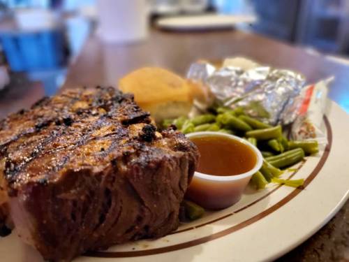 A plate featuring a grilled steak, green beans, cornbread, and a small cup of sauce.