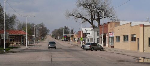 A quiet street in a small town, featuring shops, trees, and a few cars on the road under a cloudy sky.