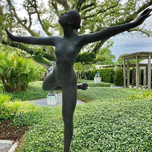 A graceful bronze sculpture of a dancer with arms outstretched, surrounded by lush greenery and other sculptures.