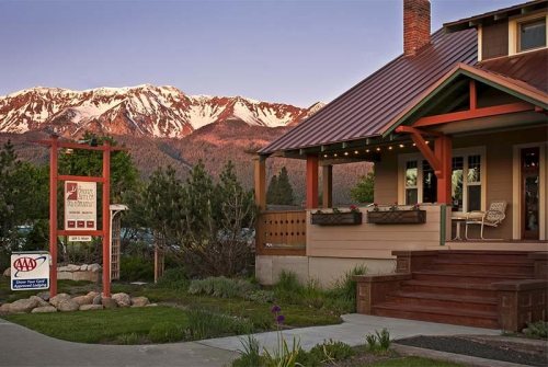 A cozy lodge with a red roof and wooden porch, set against snow-capped mountains at sunset.