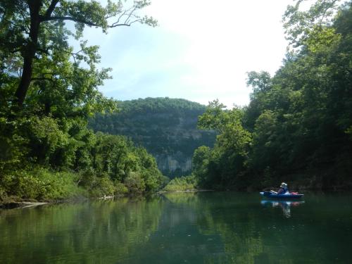 A serene river scene with lush green trees and a person kayaking in the foreground, surrounded by a mountainous landscape.