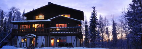 A cozy, two-story house with warm lights glowing in the windows, surrounded by snow-covered trees at dusk.