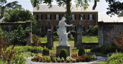 A white statue of a woman and child surrounded by colorful flowers and greenery, with a historic building in the background.