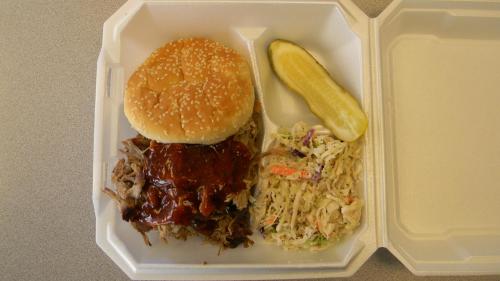 A takeout container with a pulled pork sandwich, barbecue sauce, a pickle, and a side of coleslaw.