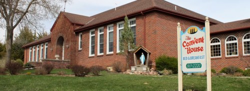 A brick building with large windows and a sign for "The Convent House Bed & Breakfast" in a landscaped setting.