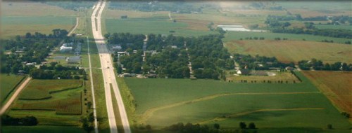 Aerial view of a highway cutting through green fields and a small town, surrounded by farmland and trees.