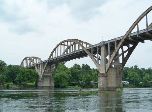 A large arch bridge spans a river, surrounded by lush greenery and a cloudy sky.