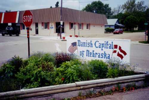 Sign reading "Danish Capital of Nebraska" in front of a small town building, with flowers and a stop sign nearby.