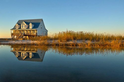 A serene house by a calm water body, reflecting the blue sky and surrounding tall grasses at sunset.