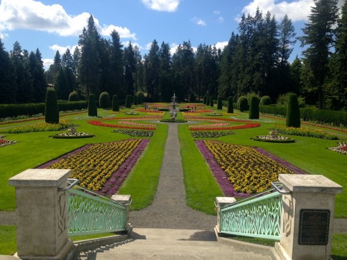 A vibrant garden with colorful flower beds, neatly arranged paths, and tall trees under a blue sky.