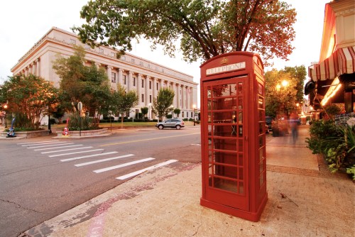 A red telephone booth stands on a sidewalk near a street, with a historic building and trees in the background at sunset.