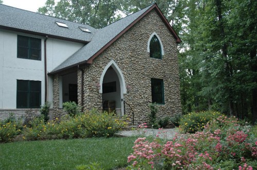 A stone-clad house with an arched entrance, surrounded by greenery and colorful flowers in a serene setting.