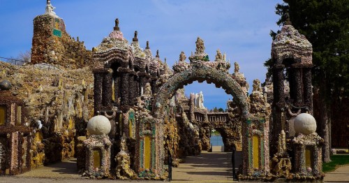 Colorful archway made of shells and stones, leading to a whimsical garden with sculptures and a blue sky above.