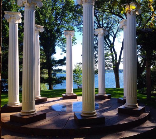 A circular stone platform with white columns overlooks a serene lake surrounded by trees.