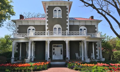 A historic two-story house with a gray facade, white trim, and a flower garden in front, under a clear blue sky.