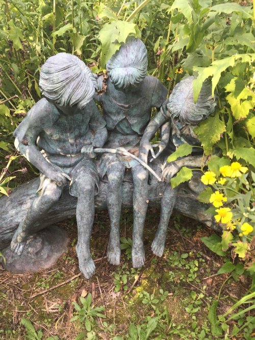 Three bronze children sitting on a log, engaged in a playful activity, surrounded by lush greenery and yellow flowers.