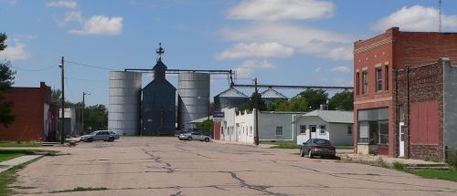 A quiet street in a small town, featuring silos in the background and a few buildings lining the road.