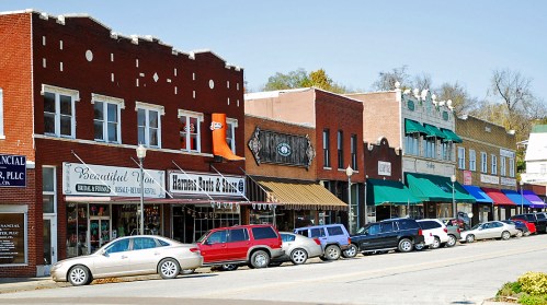 A charming street lined with shops and parked cars, showcasing a mix of brick buildings and colorful awnings.