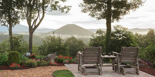 Two wooden chairs face a scenic view of mountains and misty valleys, surrounded by lush greenery and colorful flowers.