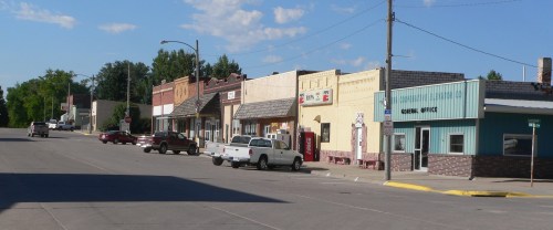 A quiet small-town street lined with storefronts and parked vehicles under a clear blue sky.