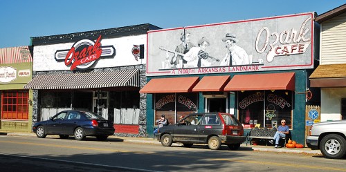 A quaint street scene featuring the Ozark Café and a nearby café, with parked cars and autumn decorations.