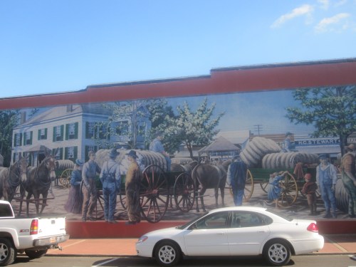Mural depicting a historical scene with people and horse-drawn wagons in a town setting, alongside a parked car.